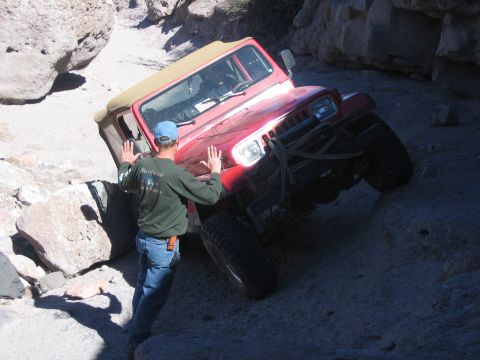 1996 Ford Bronco - A man attempts to free a red Jeep Wrangler that is stuck on a challenging, rocky off-road path.