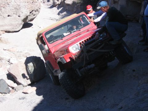 1996 Ford Bronco - A red Jeep Wrangler is being pushed by people while navigating a difficult rocky trail.