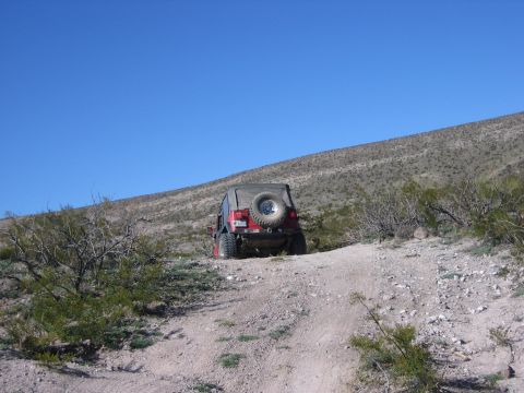 1996 Ford Bronco - A red Jeep Wrangler navigates a dusty trail in a dry, hilly terrain under a clear blue sky.