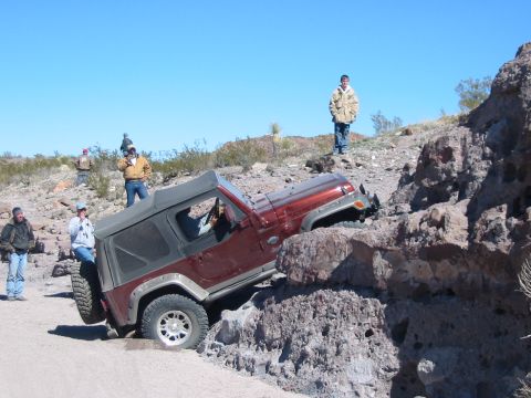 1996 Ford Bronco - A red Jeep Wrangler maneuvers through a challenging rocky obstacle on a sunny day.