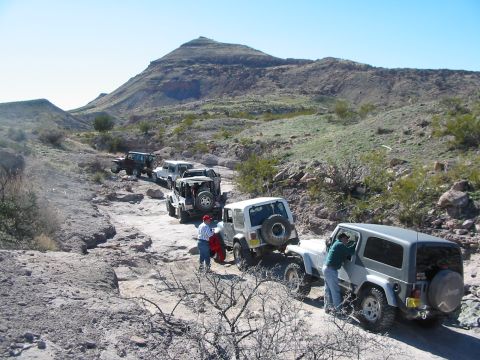 1996 Ford Bronco - A convoy of Jeeps navigates a challenging off-road path under a clear blue sky.