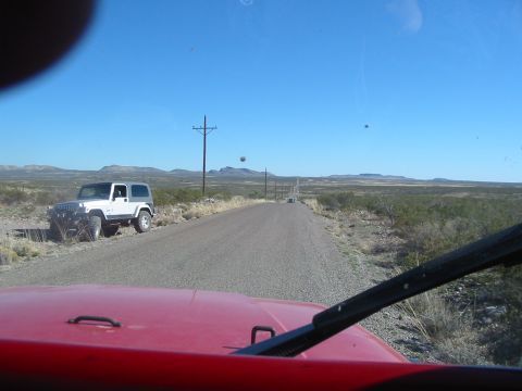 1996 Ford Bronco - View from inside a red vehicle looking out at a white Jeep parked on a dusty road with mountains in the distance.