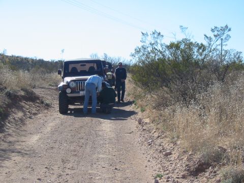 1996 Ford Bronco - People are gathered around the front of a Jeep Wrangler on a dusty trail, with another vehicle behind.