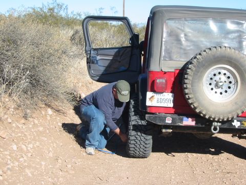 1996 Ford Bronco - A man kneels to work on the rear tire of a red Jeep Wrangler parked on a dirt road.