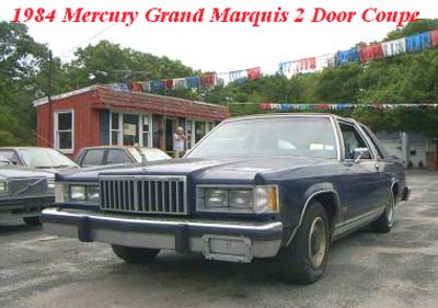 1984 Mercury Grand Marquis - A blue 1984 Mercury Grand Marquis 2-door coupe is parked in front of a building with festive banners.