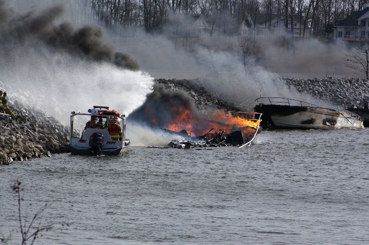 Fire department boat spraying water on a burning boat near a rock breakwall
