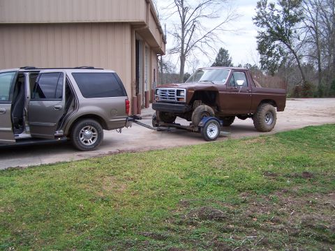 1981 Ford Bronco - A tan SUV tows a lifted brown pickup truck on a trailer in front of a building.