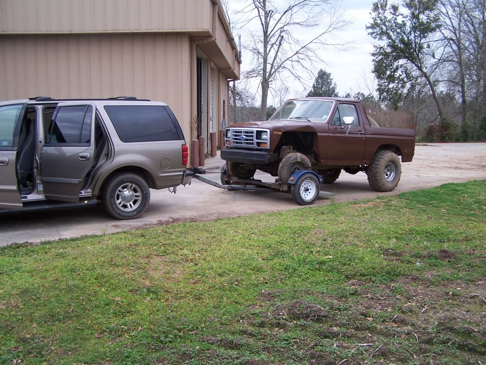 Brown pickup truck on a trailer being towed by a tan SUV. The truck has large tires and is lifted