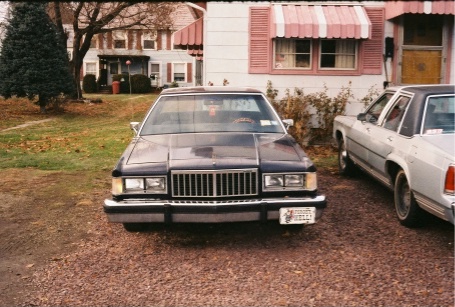 1984 Mercury Grand Marquis - A dark sedan with a prominent vertical grille is parked in a gravel driveway next to another car.