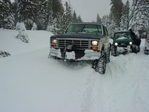1986 Ford Bronco - Two off-road vehicles navigate a snow-covered path through a forest.