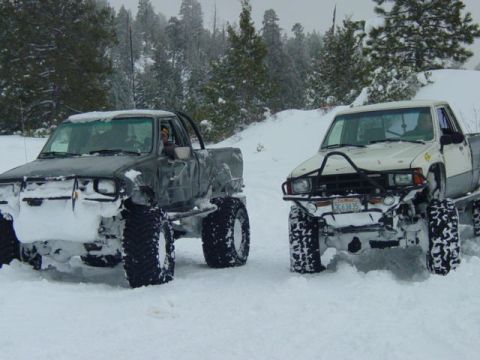 1986 Ford Bronco - Two modified pickup trucks are navigating a snowy terrain with heavy snowfall.