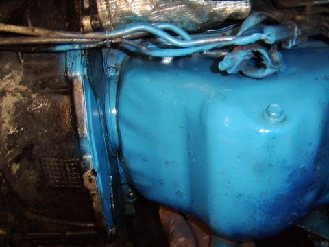 1989 Ford Bronco - Underside view of a blue-painted oil pan bolted to the engine block, with metal fuel and brake lines running above it and a hex-head bolt v…