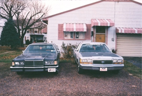 1984 Mercury Grand Marquis - Two sedans are parked in front of a house with awnings over the windows and garage.