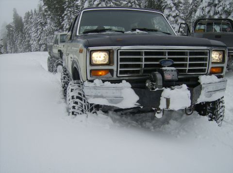 1986 Ford Bronco - A dark blue and white Ford Bronco navigates through a snowy landscape, with another vehicle visible in the background.