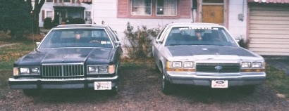 1984 Mercury Grand Marquis - Two sedans are parked side-by-side in front of a residential building with a garage door.