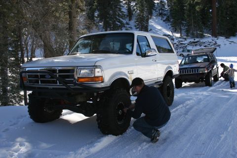 1996 Ford Bronco - A man checks the large tire of a white 1996 Ford Bronco parked in the snow, with another SUV and people in the background.