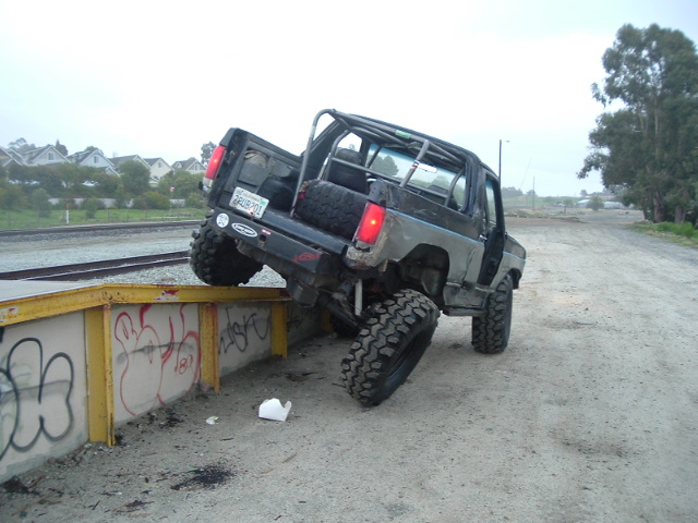 Green Ford Bronco stuck nose-down over a graffiti concrete ledge on gravel, front tires hanging off.