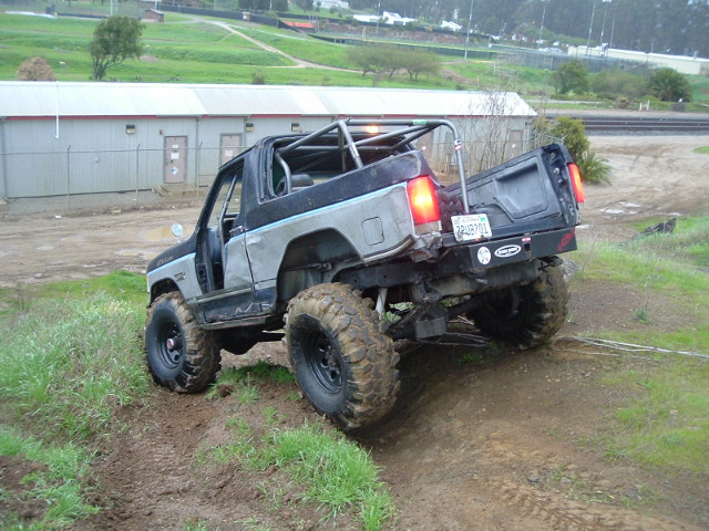 Topless gray Ford Bronco with roll cage flexing over a mud berm on 35s