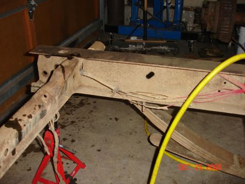 1978 Ford Bronco - The rusty frame of a vehicle is elevated on jack stands in a garage setting, with various hoses and wires attached.