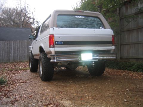 1996 Ford Bronco - Rear view of a lifted white full-size Ford Bronco with a tan hardtop, tan lower cladding and a Ford oval on the tailgate, parked on a leaf-…