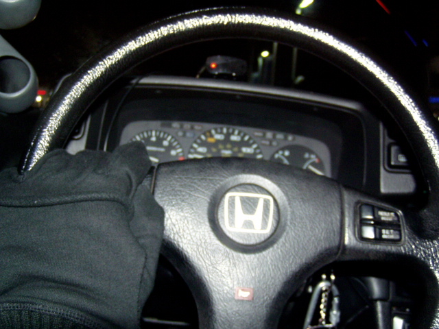 Driver's perspective inside a car, showing the steering wheel with a Honda logo and the dashboard gauges