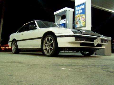 1993 Ford Probe - A white coupe with aftermarket wheels and a black hood is parked at a gas station at night.