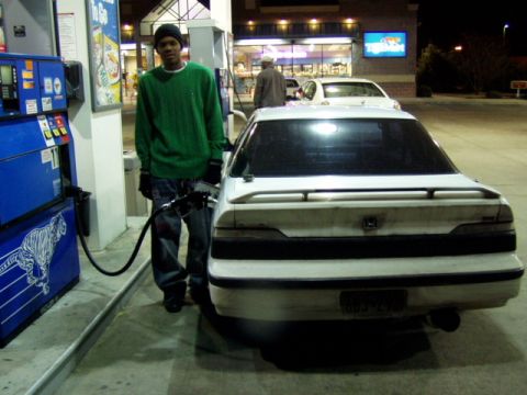 1993 Ford Probe - A person pumps gas into a white sedan at a brightly lit gas station after dark.