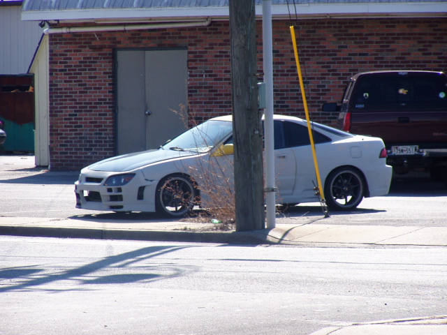 White coupe with a custom body kit parked next to a utility pole. A dark pickup truck is visible behind it