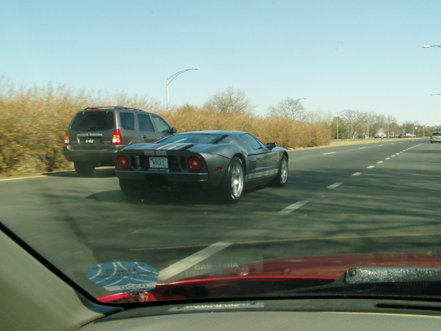 Dark grey Ford GT with white racing stripes on rear, driving on highway next to SUV. License plate reads 'RICES'