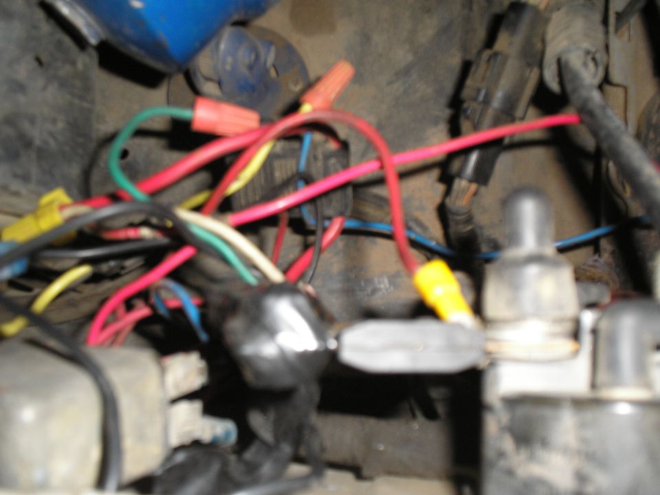 Close-up of a messy, tangled wiring harness with red, yellow, green, and blue wires connected by wire nuts and electrical tape