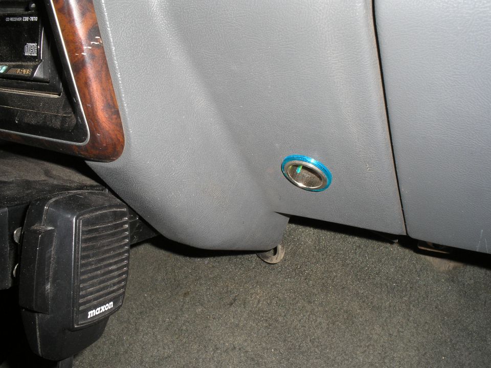 Gray interior trim panel with a blue-rimmed grommet hole near the pedal