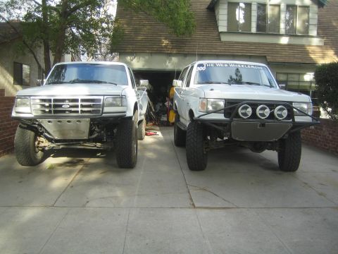 1990 Ford Bronco - Two white Ford prerunner pickups sit side by side on a driveway in front of a house, one with triple round lights.
