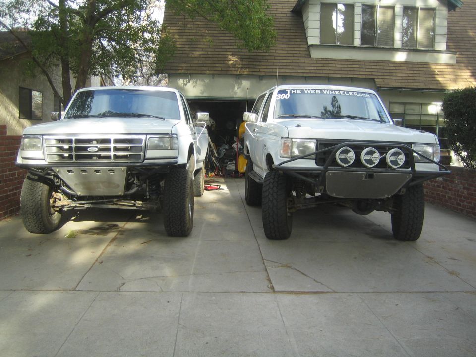 Two white Ford prerunner trucks parked side by side on a residential driveway