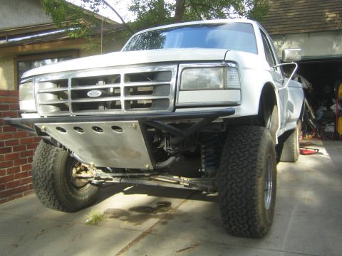 1990 Ford Bronco - white Ford prerunner truck shows its front long-travel suspension and skid plate while parked on a driveway by a garage.