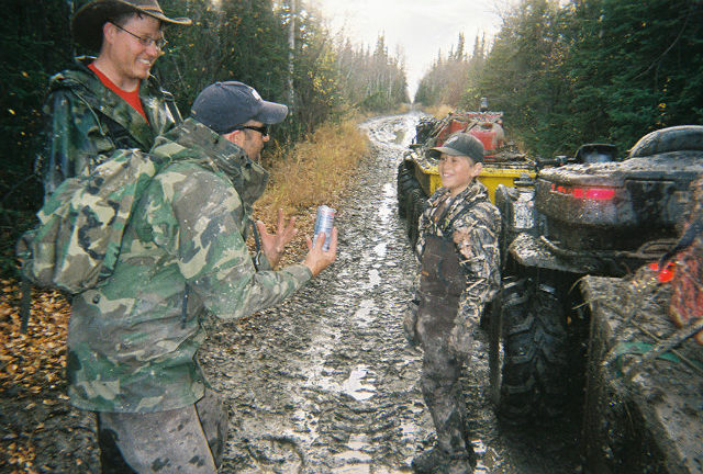 Two muddy ATVs on a rutted forest trail with two adults and a boy in camouflage clothing