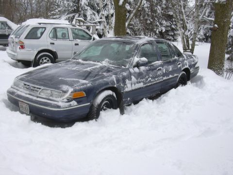 1996 Ford Crown Victoria - A dark blue 1996 Ford Crown Victoria is partially buried in snow, with another vehicle visible in the background.