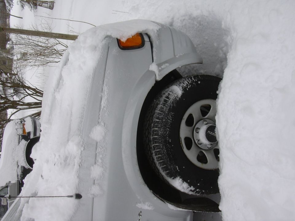 White truck buried in snow, only front corner and wheel visible. Another vehicle is partially visible to the left