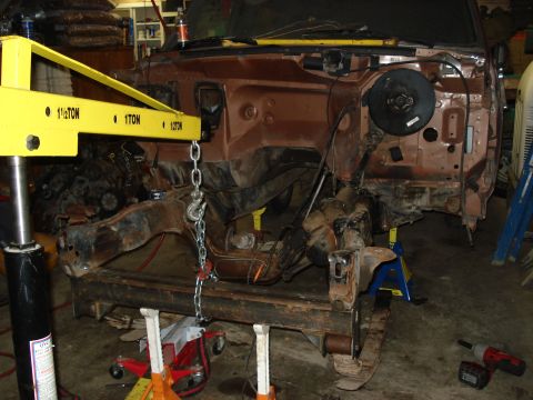1989 Ford Bronco - The vehicle's front end is disassembled, with the chassis supported by jack stands and a yellow engine hoist.