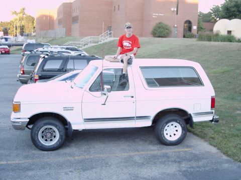 1988 Ford Bronco - A person sits atop a white Ford Bronco in a parking lot, with other vehicles and a brick building in the background.