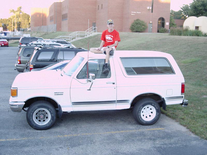 guy sitting on roof of white Ford Bronco, parked next to other vehicles