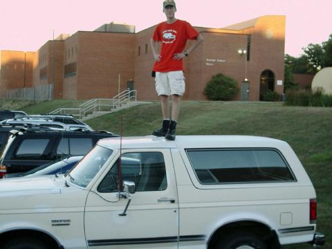 1988 Ford Bronco - A man stands atop a white SUV parked in front of a brick building.