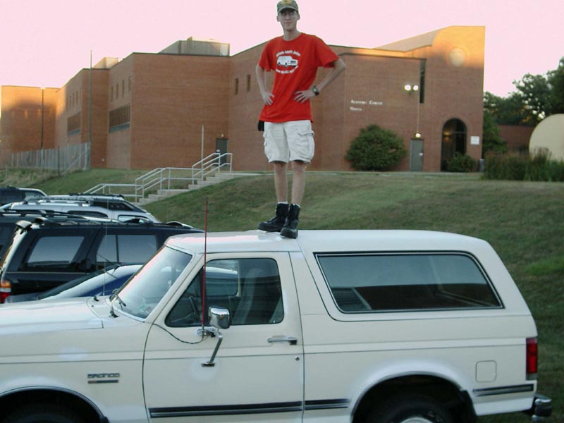 White SUV with a man standing on its roof, side view. The vehicle has a "BRONCO" badge