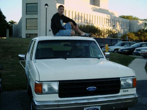 1988 Ford Bronco - A man sits atop a white Ford pickup truck in a parking lot with other vehicles and a building in the background.