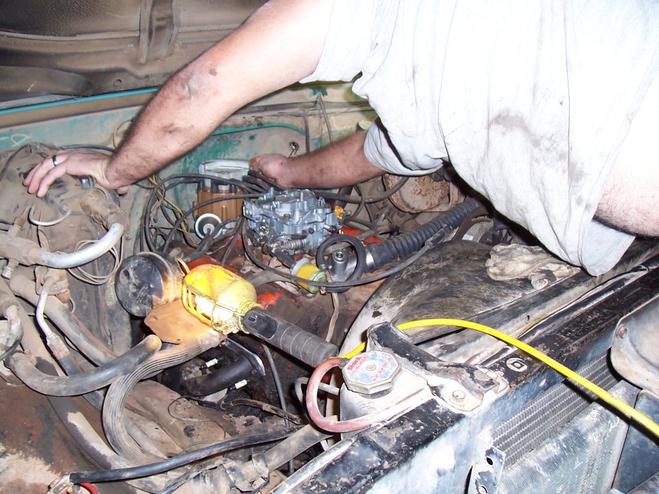 Mechanic working on a dirty engine bay, focusing on the carburetor. A work light illuminates the area