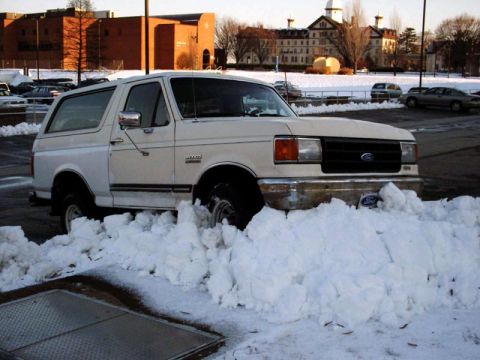 1988 Ford Bronco - A white Ford Bronco is stuck in deep snow in a parking lot with buildings in the background.
