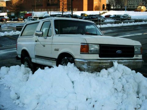 1988 Ford Bronco - A white Ford Bronco is partially buried in a large snowdrift, its front end facing the camera.