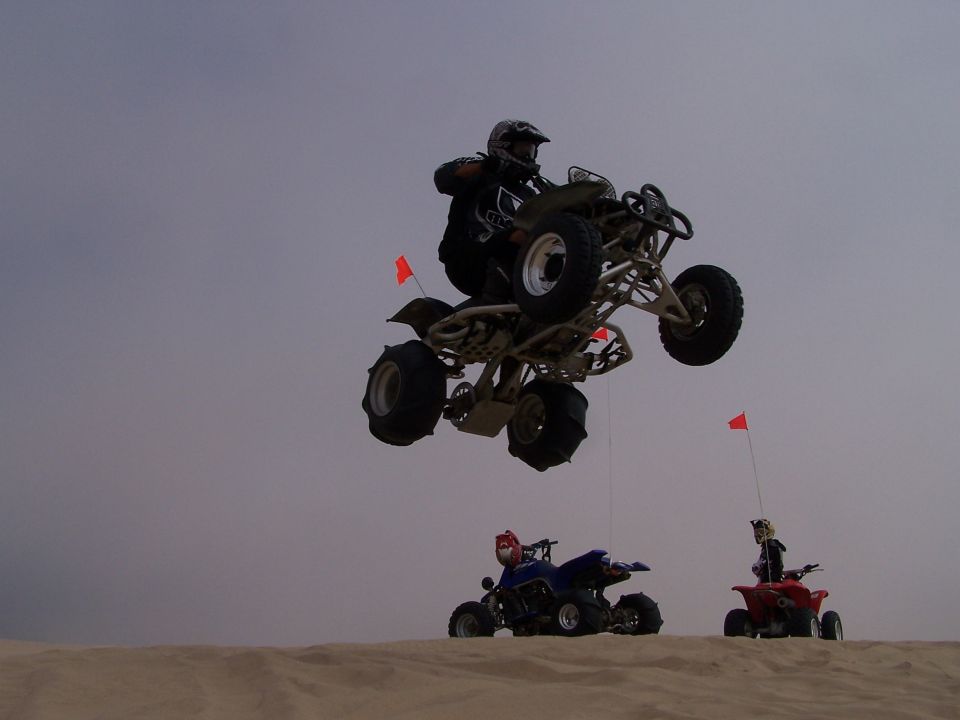 Three ATVs airborne over sand dunes under a hazy sky