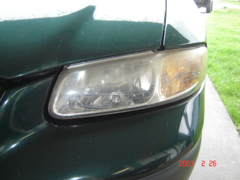 Close-up of headlight on a dark green car with sidewalk in background