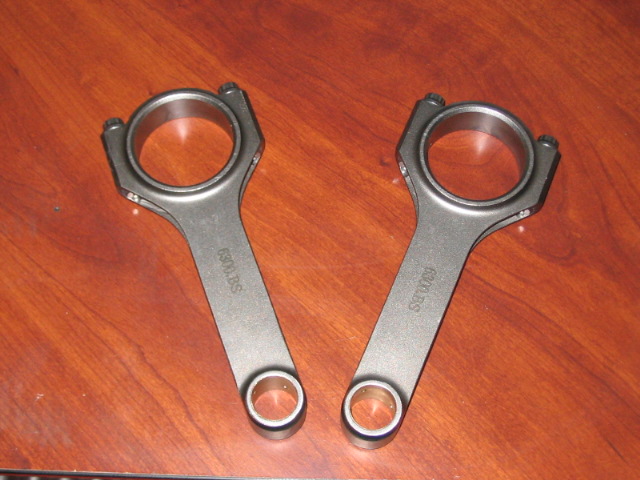 Two connecting rods with "6300.BS" stamped on them sit on a wooden surface
