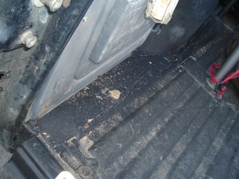 1989 Ford Bronco - The interior floor of a vehicle shows black carpeting and a gray plastic trim piece, with visible dirt and debris.
